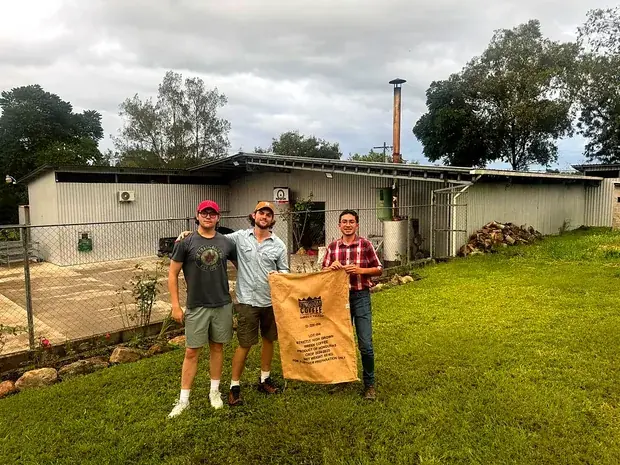 Three men holding a burlap sack of green coffee outside the mill